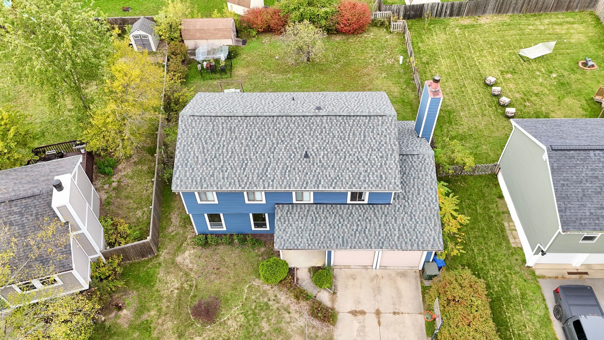 Overhead view of a blue house with gray roof, chimney, and two-car garage. Houses and greenery surround.