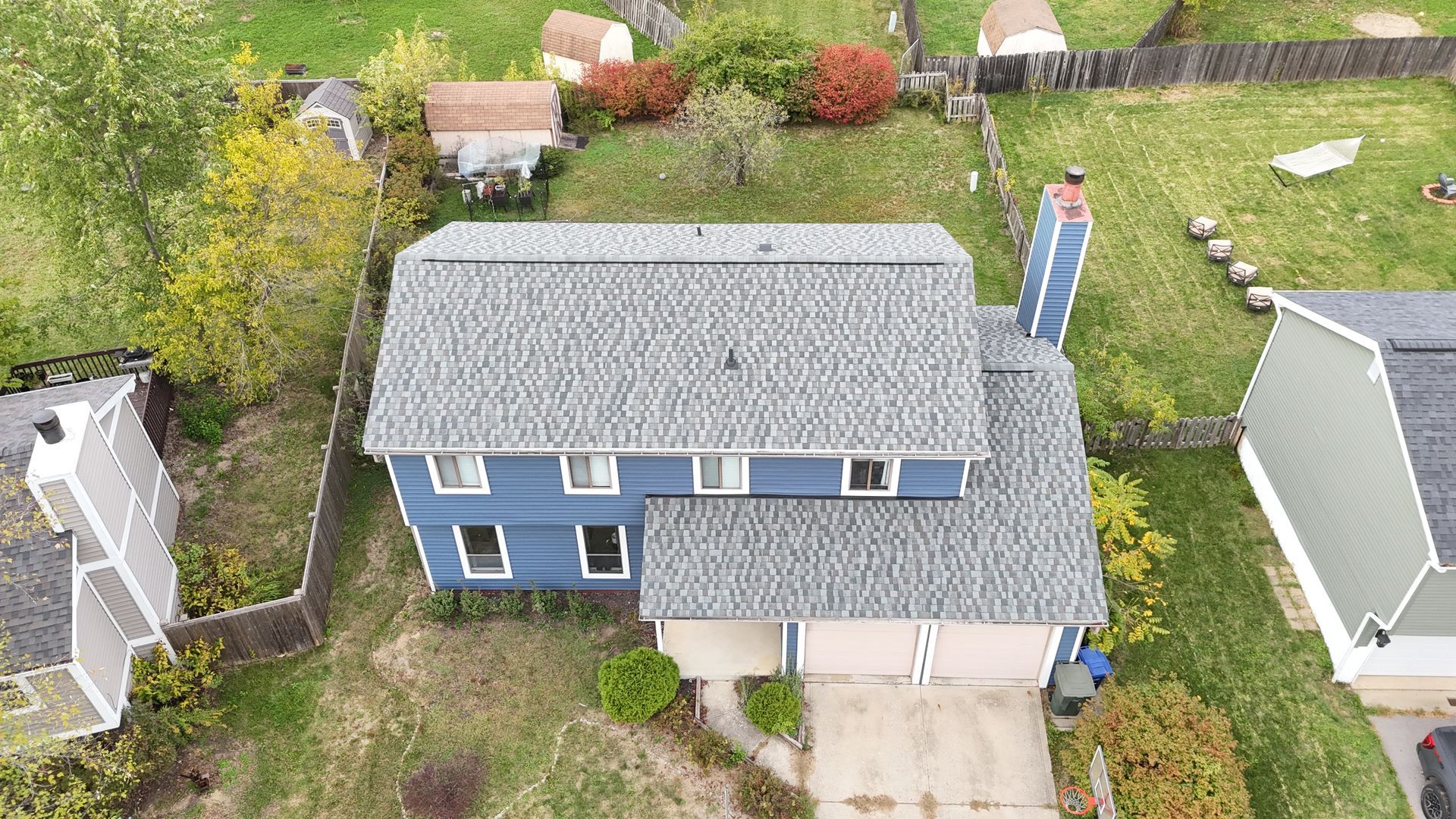 Aerial view of a blue house with gray roof, chimney, and two-car garage. Houses and trees surround it.