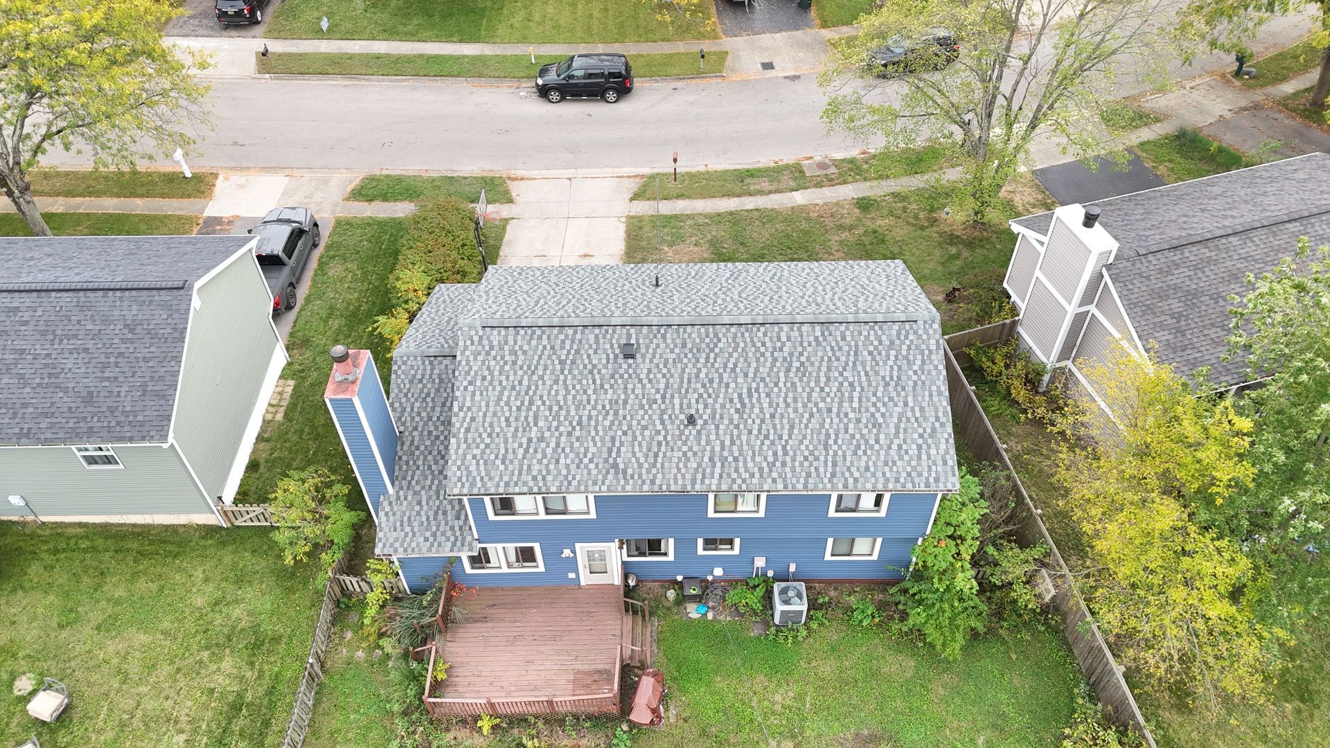 Blue two-story house with gray roof, wooden deck, chimney. View from above, residential neighborhood.