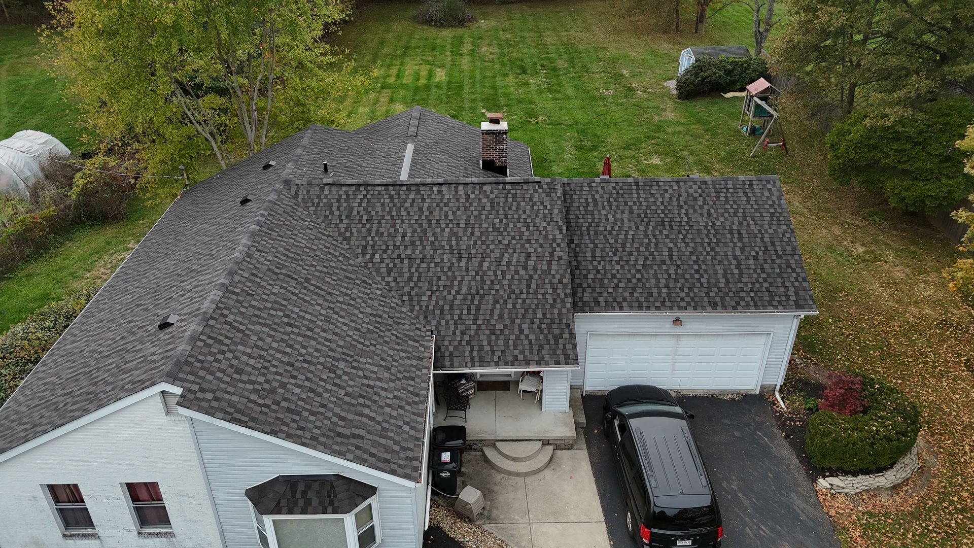 Aerial view of a house with a dark gray shingled roof, a driveway with a black van, and a green lawn.