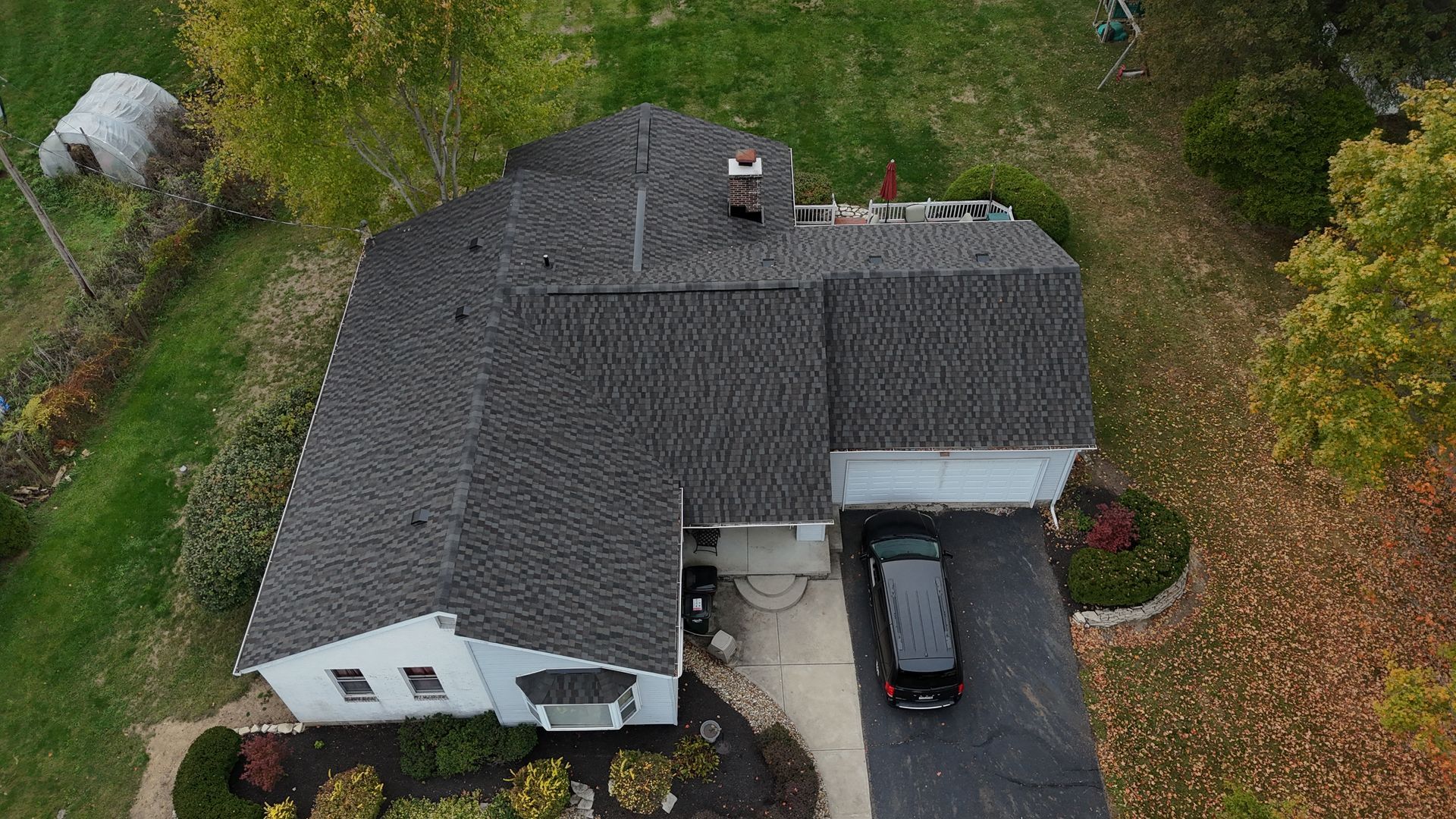 Aerial view of a gray-roofed house with a black car in the driveway, surrounded by greenery and trees.