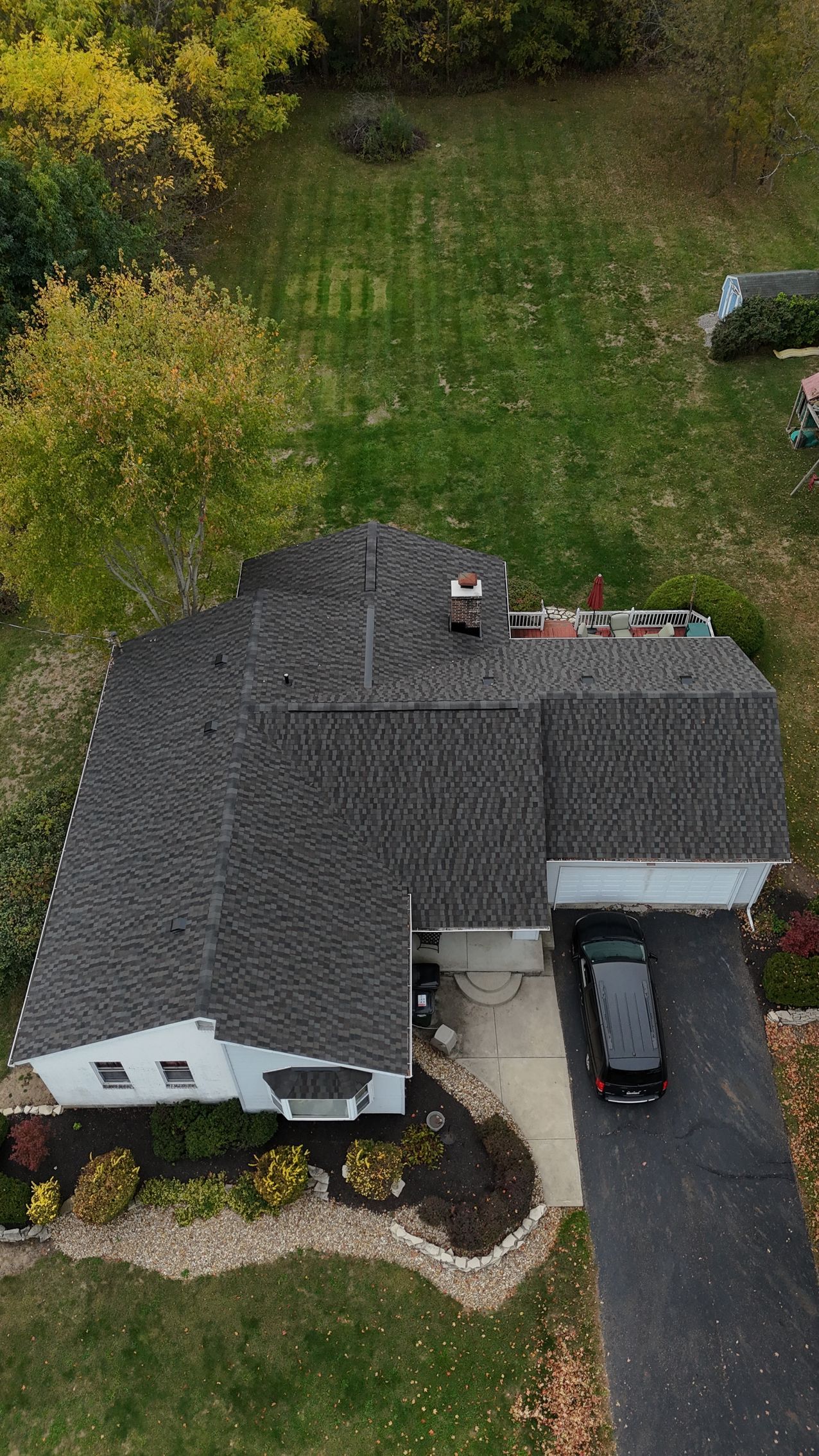 Aerial view of a house with a dark roof, driveway, and a black vehicle parked in front of a garage.