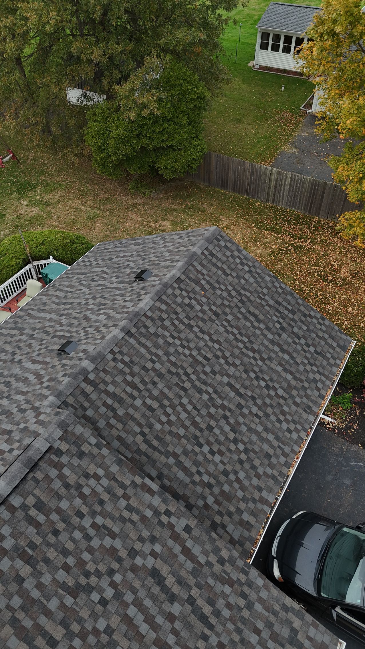Dark gray asphalt shingle roof. Green and yellow foliage in background. White gazebo and car visible.
