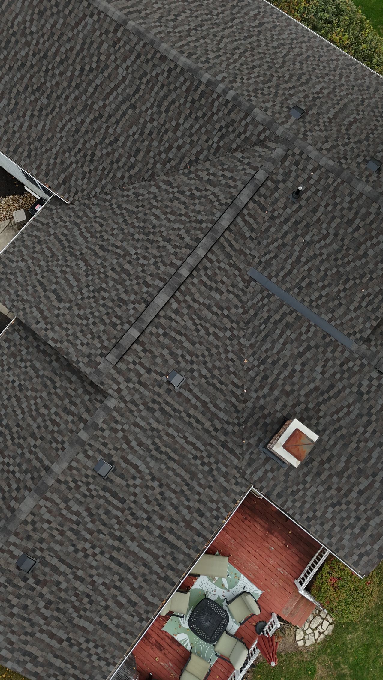 Aerial view of a house roof, covered in dark asphalt shingles, with a chimney and small porch visible.