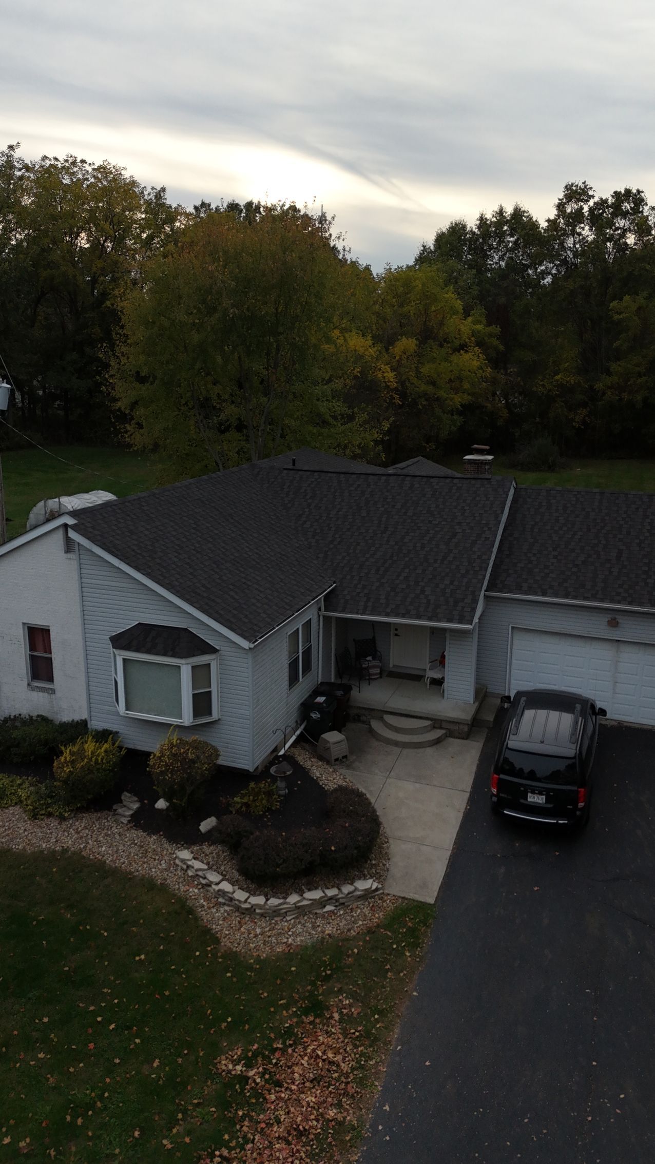 White house with black roof, bay window, driveway, car, and autumn trees.