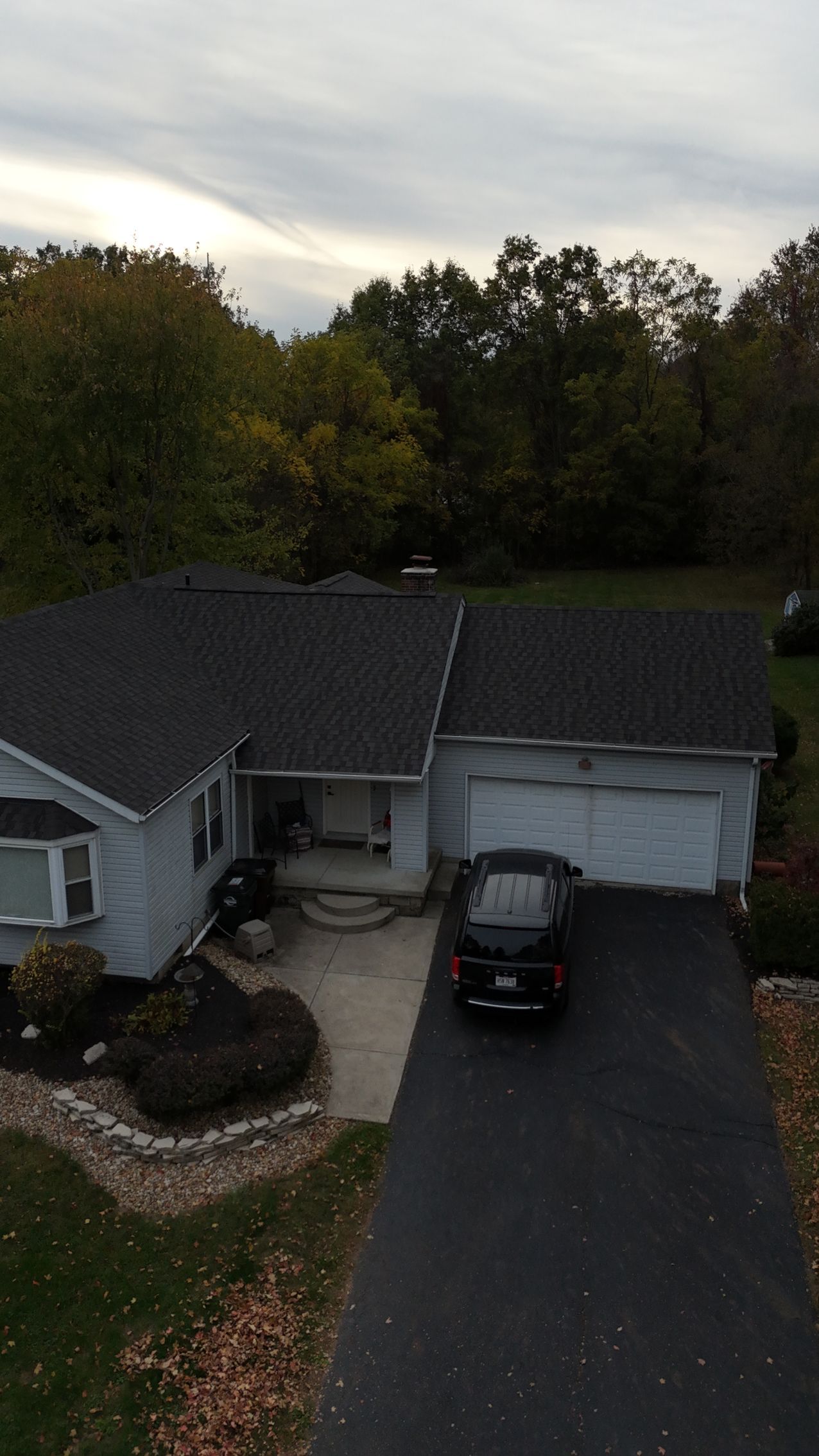 House with dark roof, black car in driveway, surrounded by trees under cloudy sky.