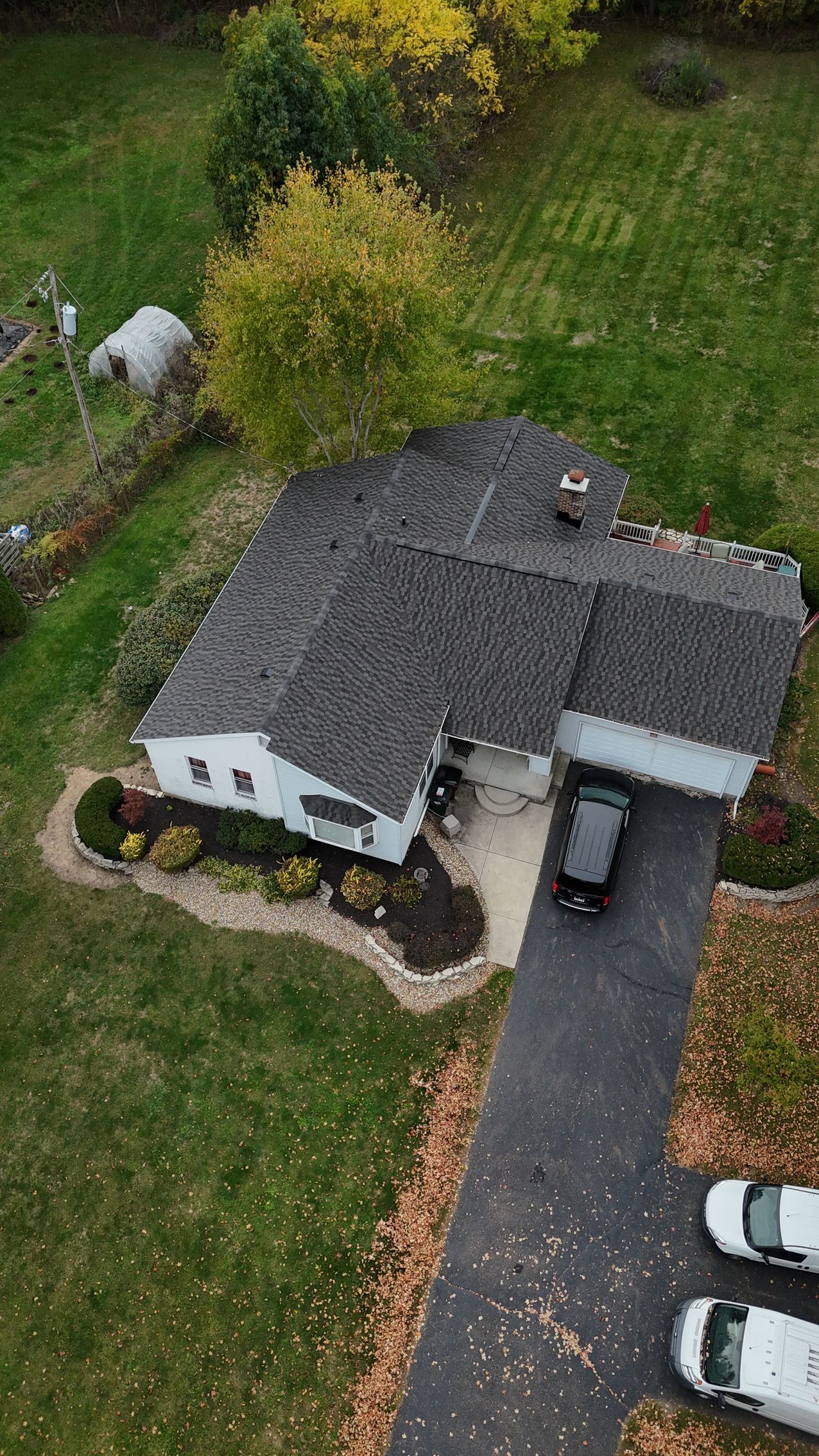 House with dark roof and driveway, surrounded by green grass and trees. Cars parked on the driveway.
