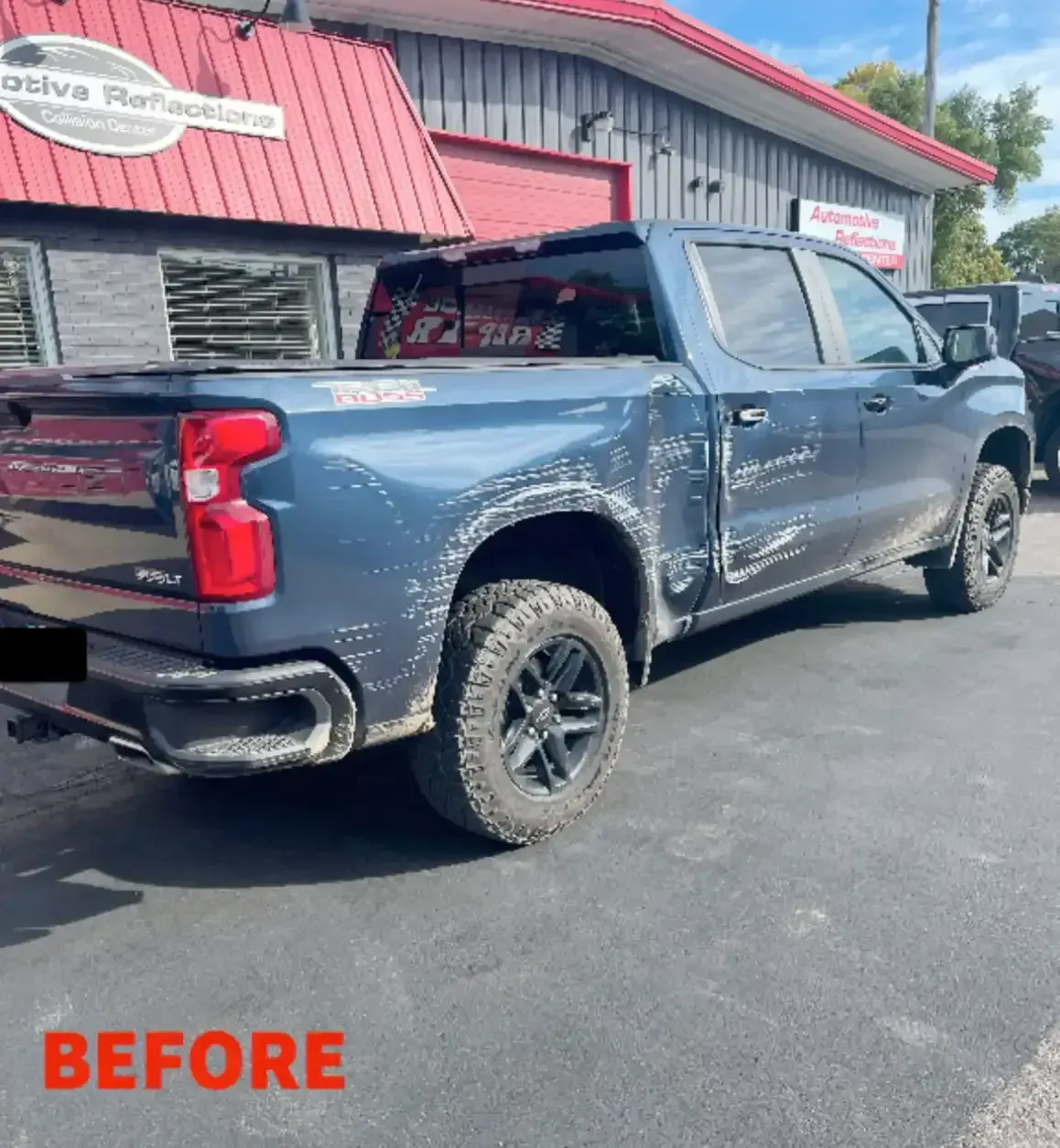 A blue pickup truck parked outside an auto body shop, showing significant dents and scratches on the side panels.