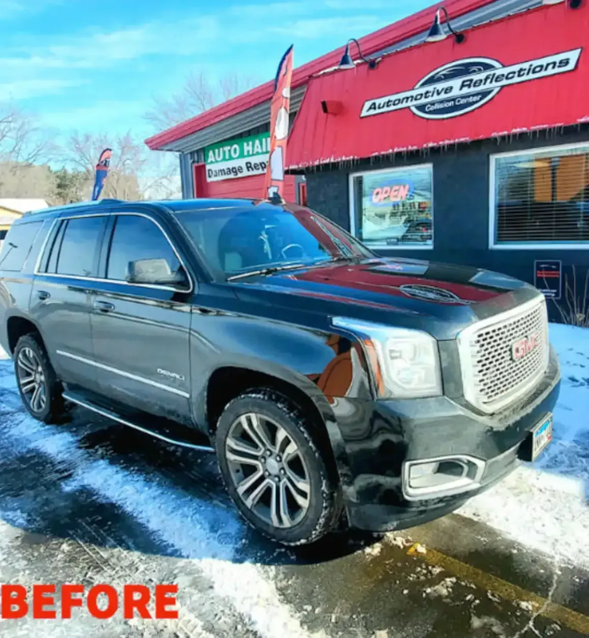 A black GMC Yukon SUV parked in front of an Automotive Reflections shop on a sunny day with snow on the ground.