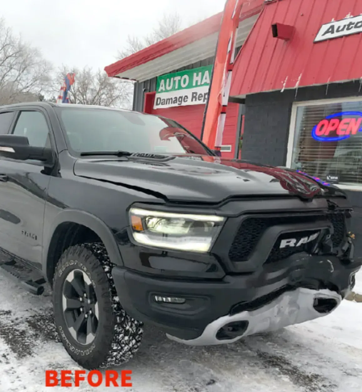 A black Ram truck with significant front-end collision damage parked in front of an auto repair shop.