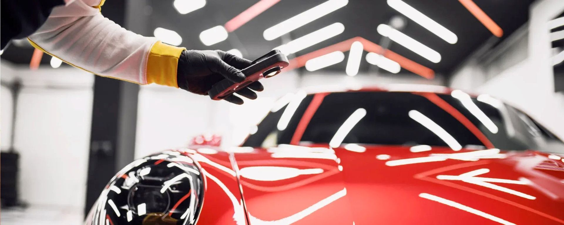 A person in black gloves uses a diagnostic light to inspect the glossy red paint of a sports car in a professional garage.