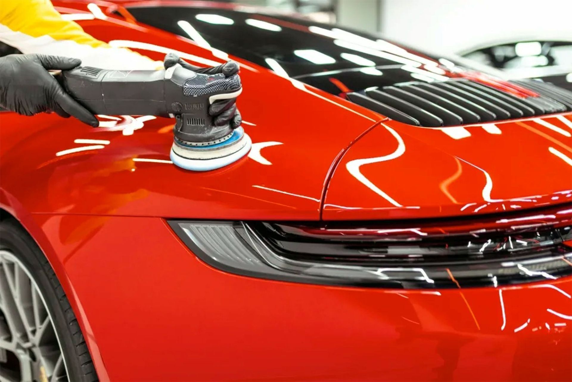 A gloved hand uses a power polisher to detail the glossy, red paint on the rear quarter panel of a sports car.