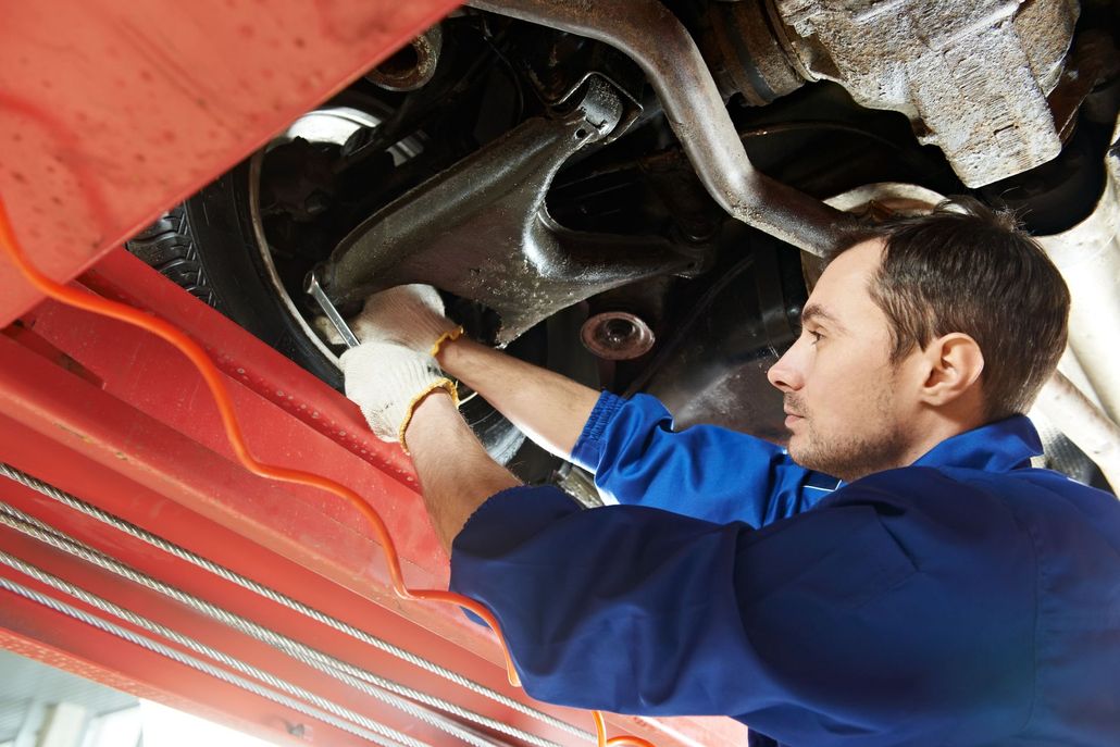 A mechanic in blue coveralls and white gloves repairs the undercarriage of a vehicle lifted on a hydraulic hoist.