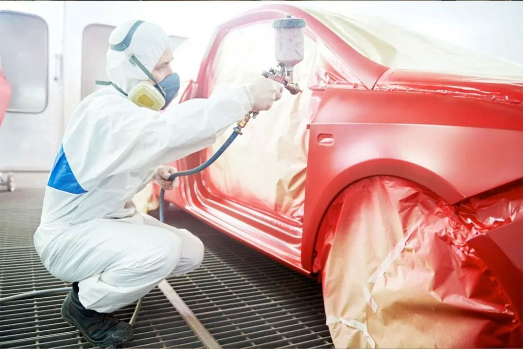 A technician in a protective suit spray-paints a red car in a professional paint booth.