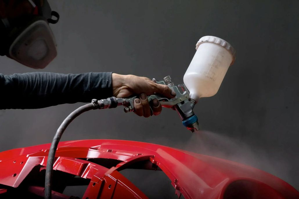 A person wearing a respirator uses a spray gun to apply a red finish to a car bumper.