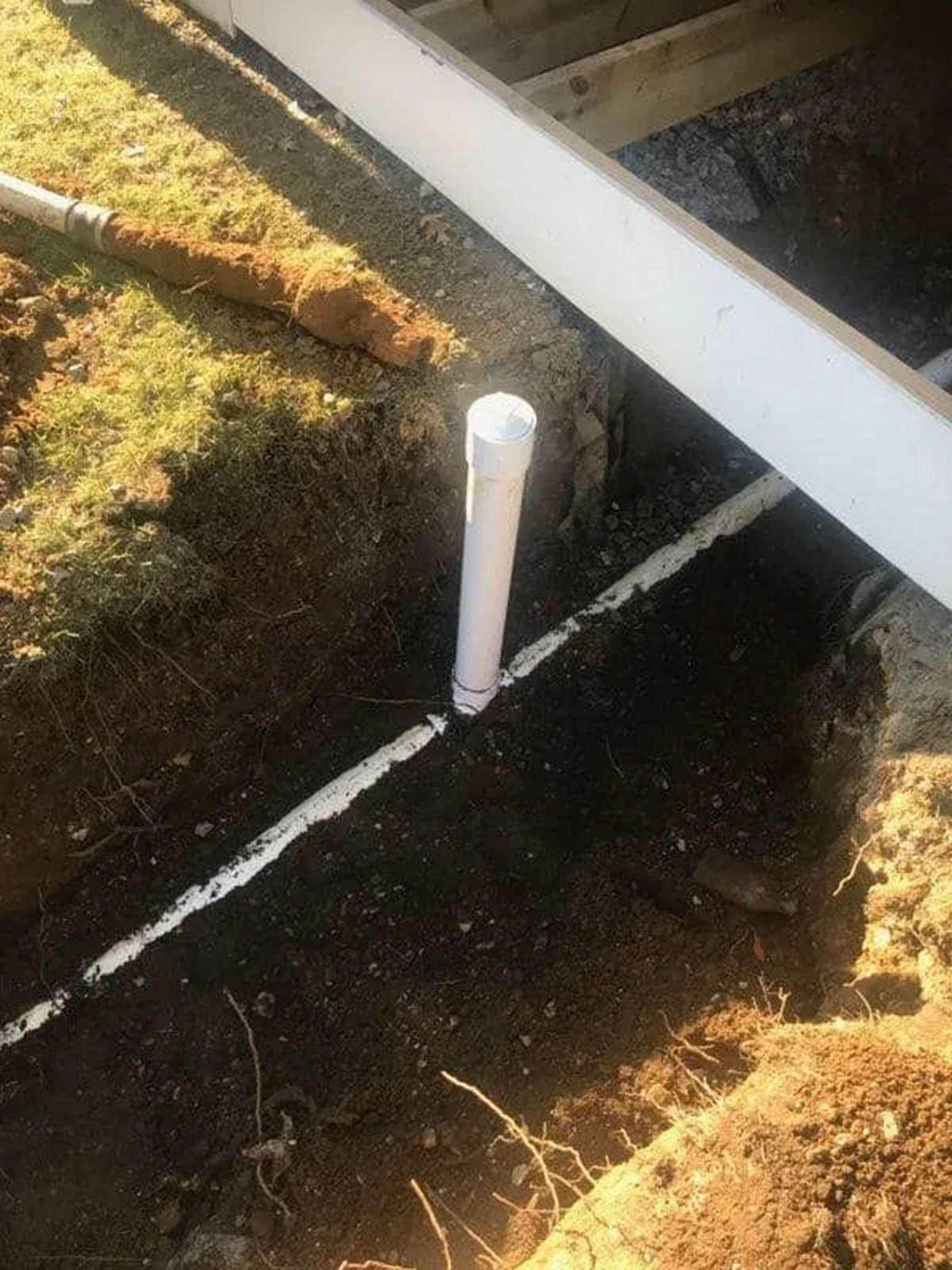 White PVC pipe emerging from dark soil beside a concrete edge and grassy area