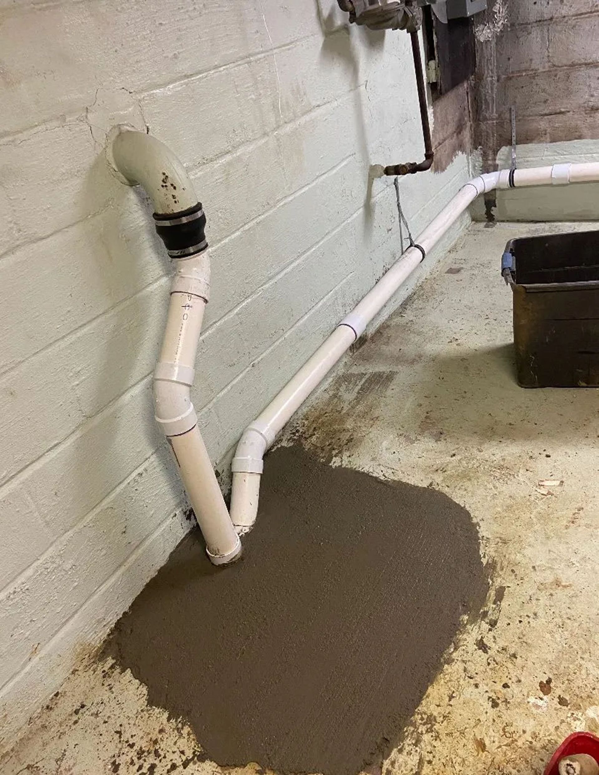 Basement wall with white pipes and a wet concrete floor around a dark puddle.