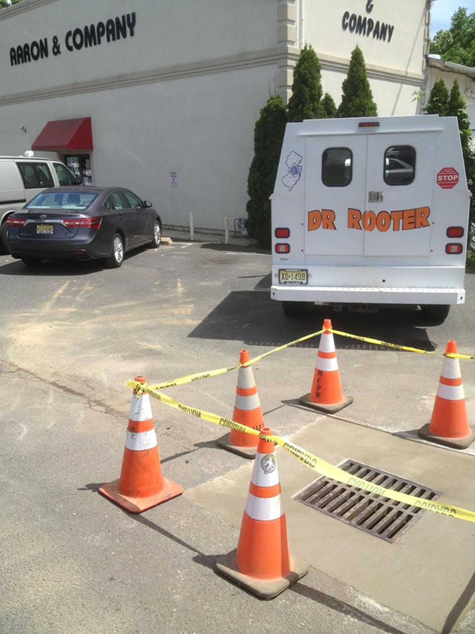 White van and black car parked by a building, with orange cones and caution tape blocking a drain area.