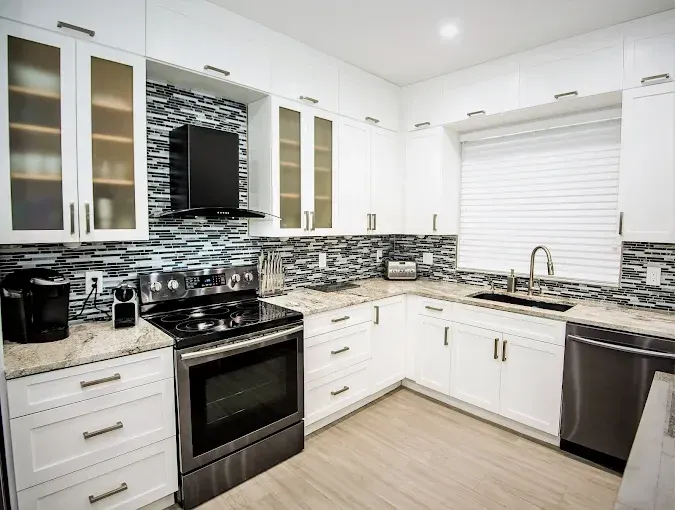 White modern kitchen with granite countertops, stainless steel appliances, and a mosaic backsplash.