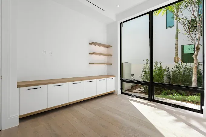 Modern office with built-in white cabinets, wooden shelves, and large window overlooking a small garden.