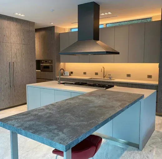 Modern kitchen with island, gray cabinets, black range hood, and a red stool.