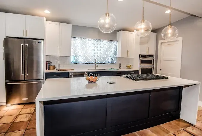 Modern kitchen with island, stainless steel fridge, white and navy cabinets, and globe pendant lights.
