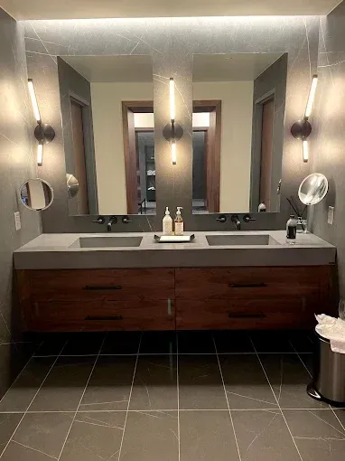 Modern bathroom with double sinks, wood cabinets, grey countertops, large mirrors, and vertical sconces.