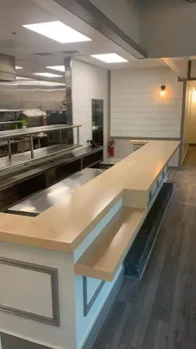 Empty restaurant bar with light wood countertop, stainless steel and white accents, and wooden floor.