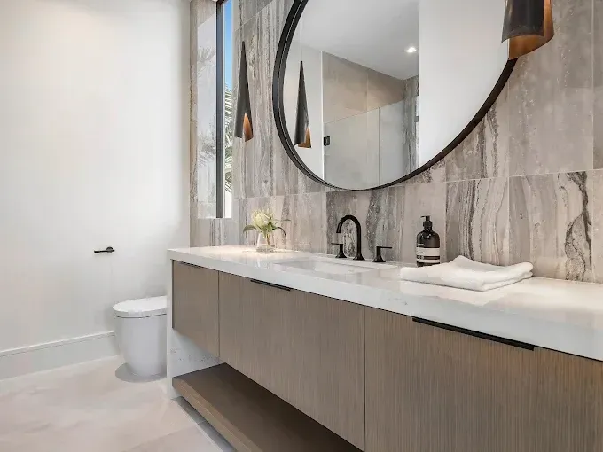 Modern bathroom with a wood vanity, marble walls, round mirror, and black fixtures.