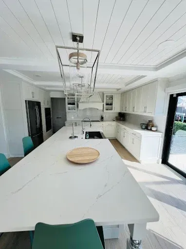 Modern white kitchen with long island, teal chairs, stainless steel appliances, and wood flooring.