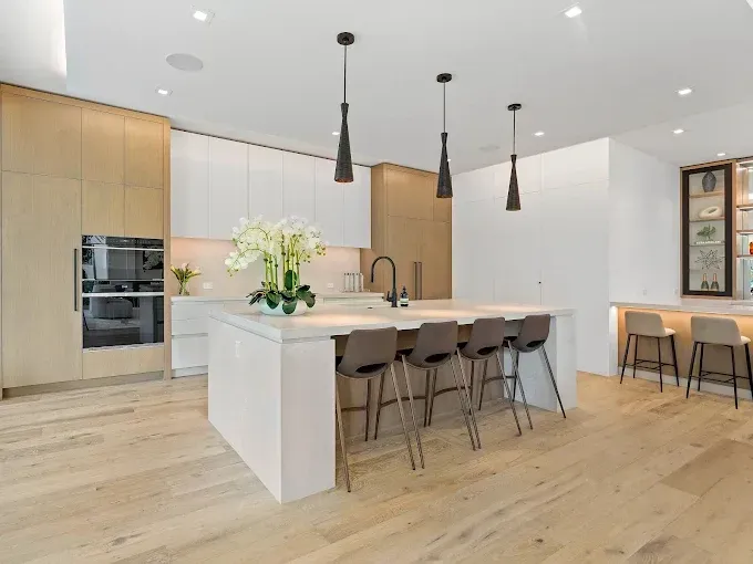 Modern kitchen with a white island, light wood floors, and three black pendant lights.