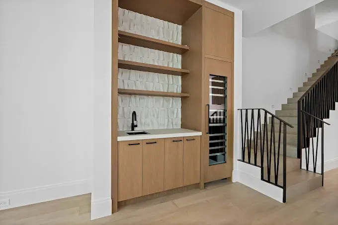 Built-in wooden bar with shelves, cabinets, sink, and wine cooler, next to a staircase with a metal railing.