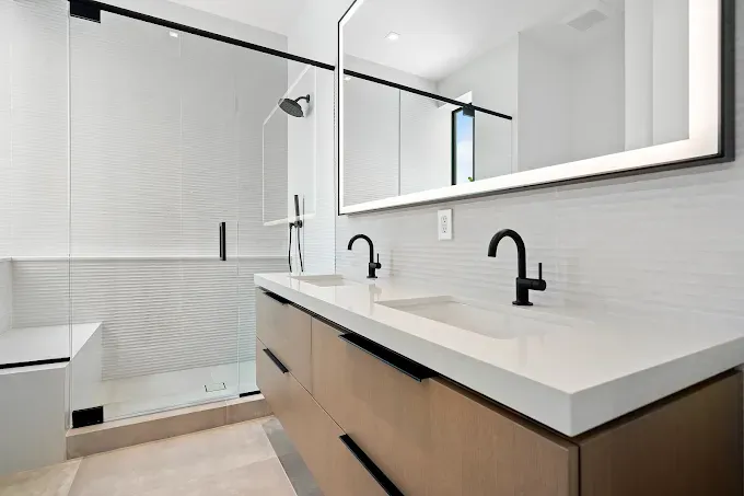 Modern bathroom with double vanity, shower, and large mirror. Light wood cabinets, white countertop, black fixtures.
