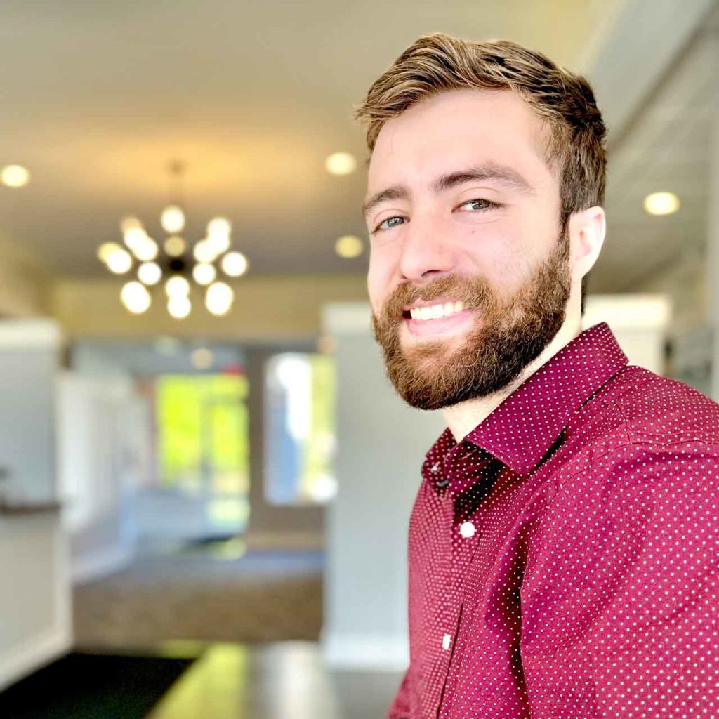 A man with a beard is wearing a red polka dot shirt and smiling.