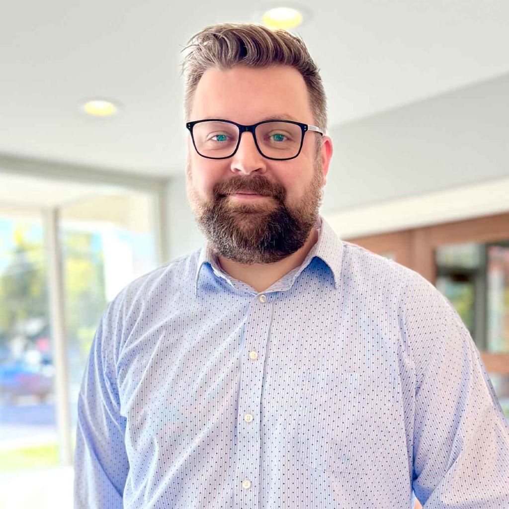 A man with a beard and glasses is standing in front of a window.