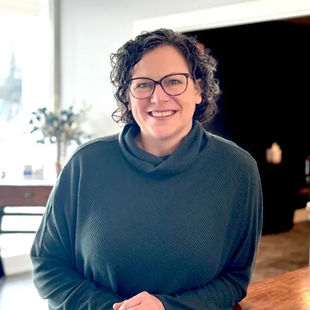 A dark haired woman with glasses with a blue top smiling