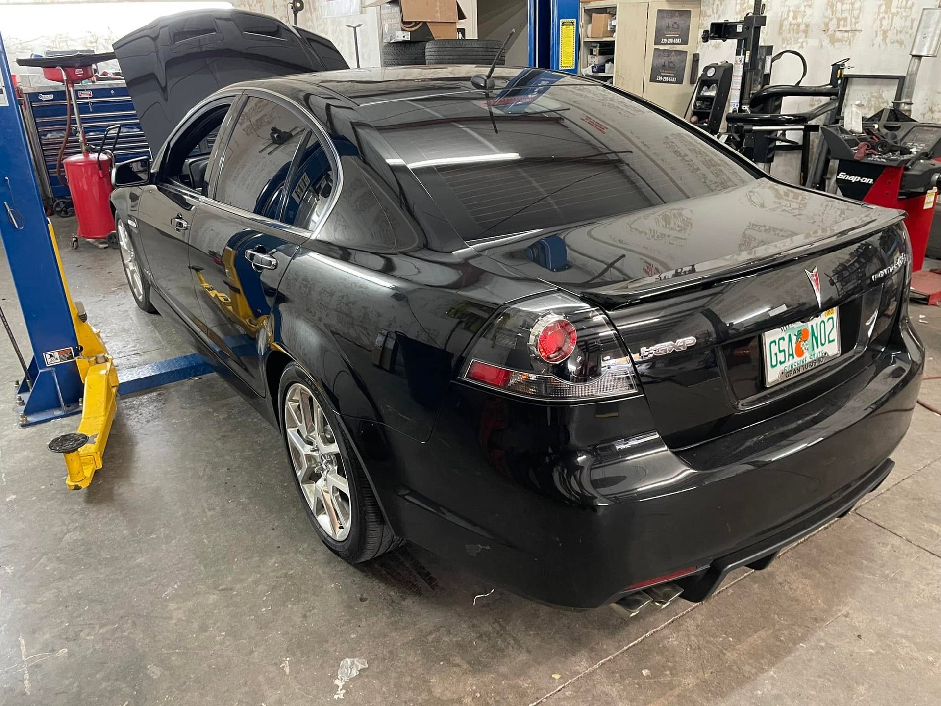 Black sedan in an auto shop with its hood open, viewed from the rear.