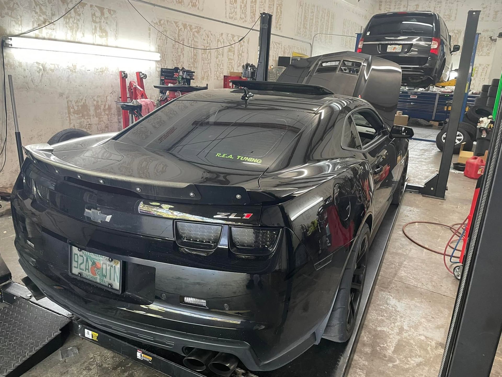 Black sports car in a garage wash bay, viewed from the rear with another car on a lift behind it