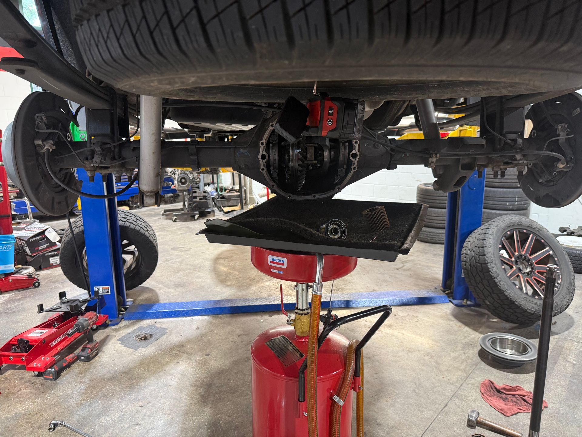 Car on a lift in a repair shop, with a red transmission jack supporting the underside.