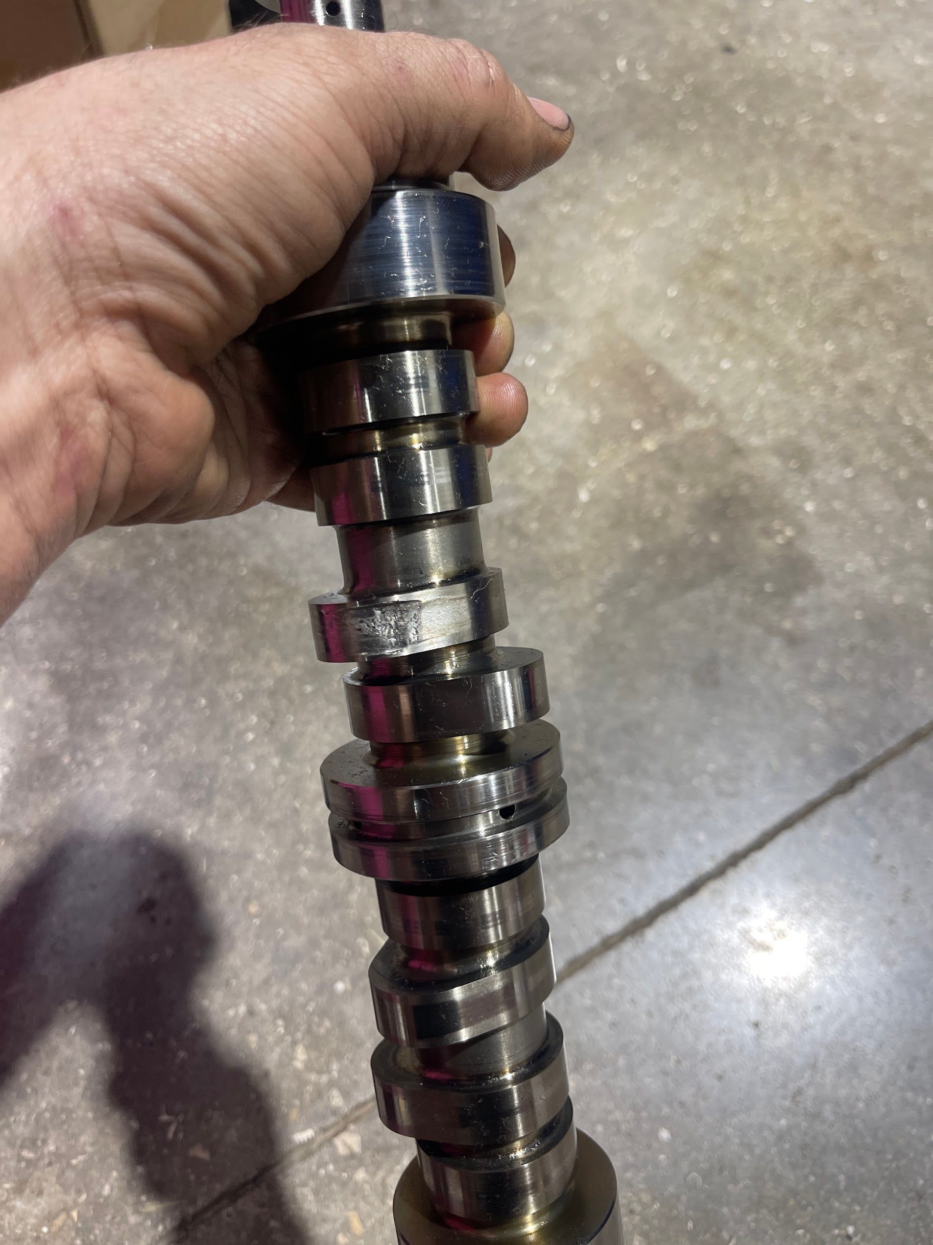 Hand holding a shiny metal camshaft or engine shaft with multiple lobes against a concrete floor.