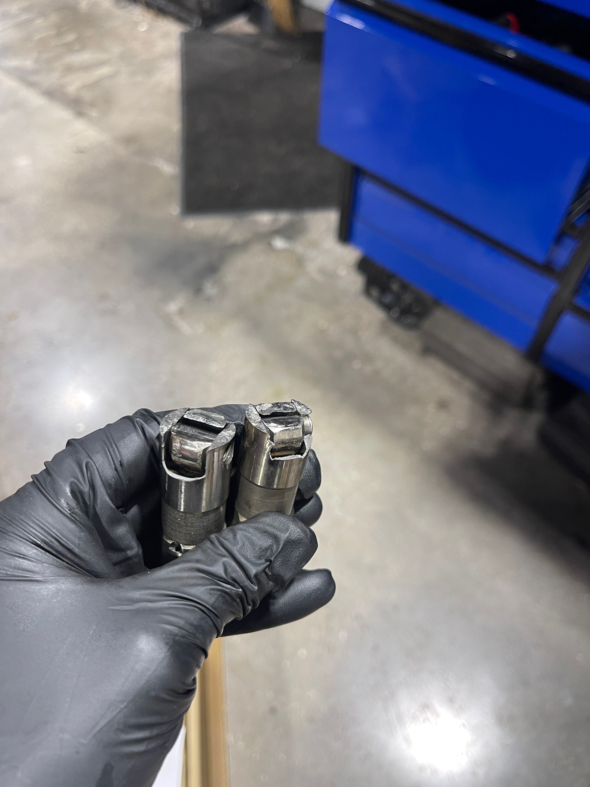 Gloved hand holding two worn metal cylinders in a workshop beside a blue bin.