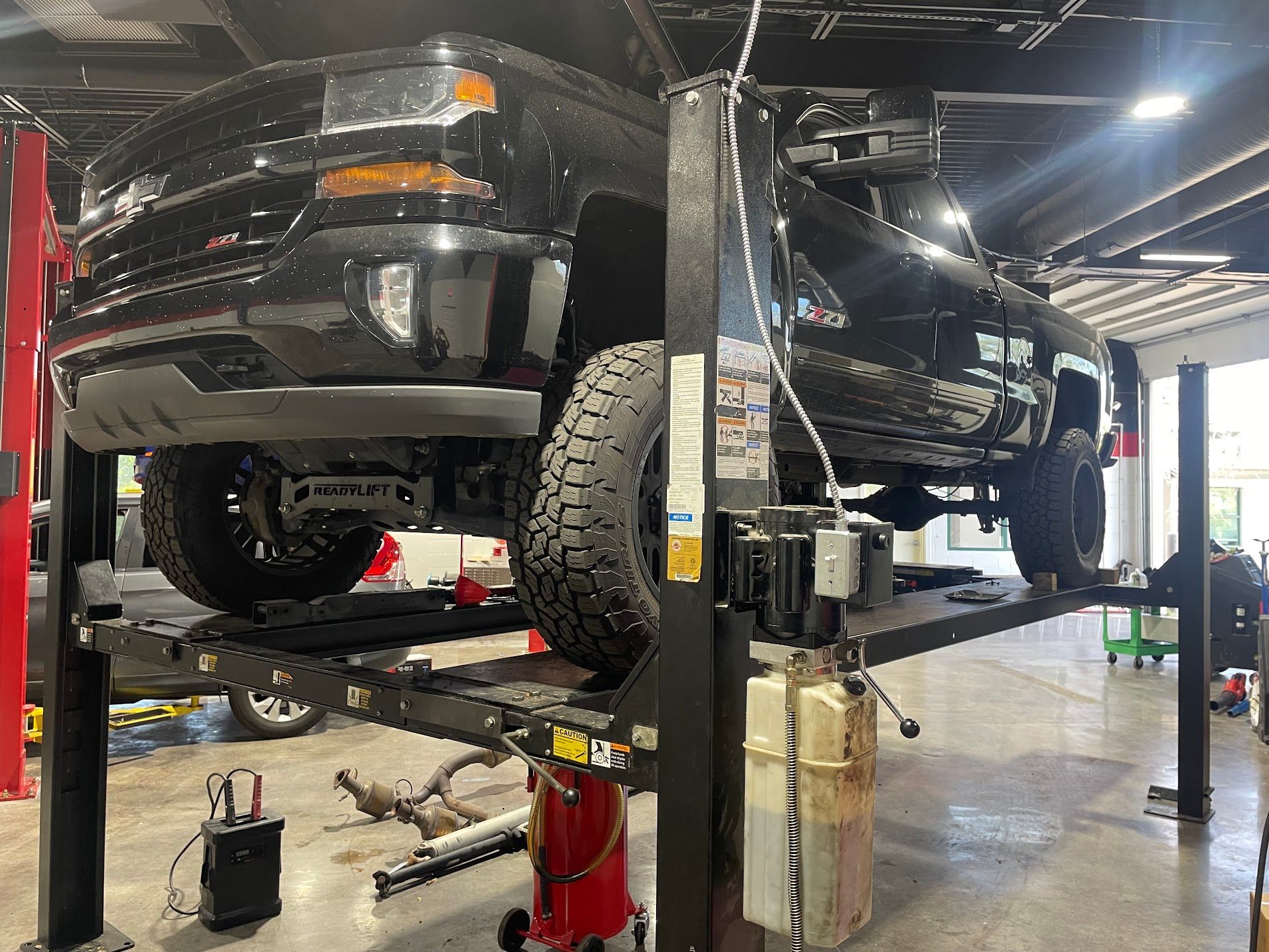 Black pickup truck lifted on a service hoist in an auto repair shop, with tools on the floor.