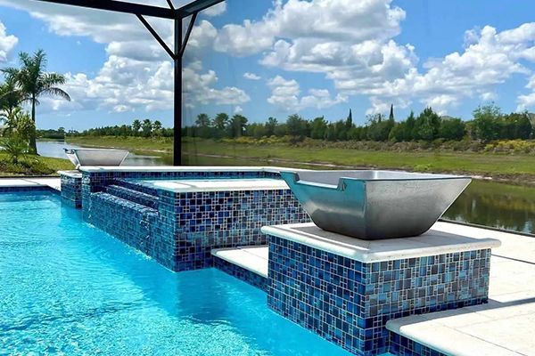 Modern blue-tiled pool with a raised spa and bowl fountain under a covered patio, overlooking a green landscape