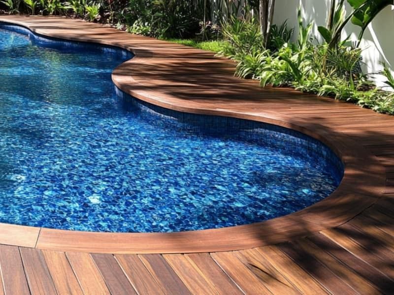 Curved blue swimming pool beside a wooden deck and lush tropical plants