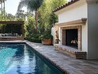 Backyard pool beside a white stucco fireplace and stone patio, with lush greenery and palm trees.