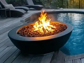 Outdoor fire pit beside a blue pool at dusk, with flames burning in a black stone basin