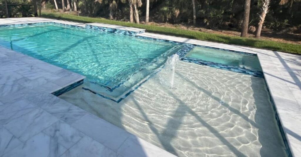 Modern backyard pool with aqua water, shallow lounging shelf, and marble-tile deck in sunlight