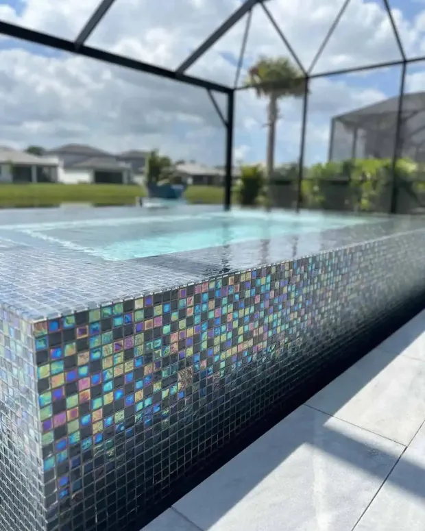 Colorful mosaic-tiled pool edge beside clear water under a screened patio with palm trees beyond