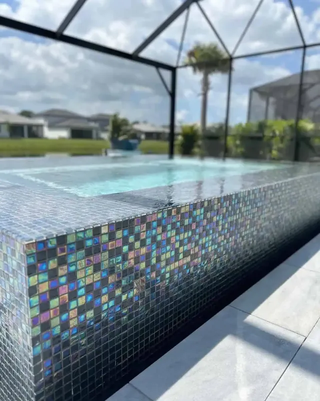 Colorful mosaic-tiled pool edge beside clear water under a screened patio with palm trees beyond