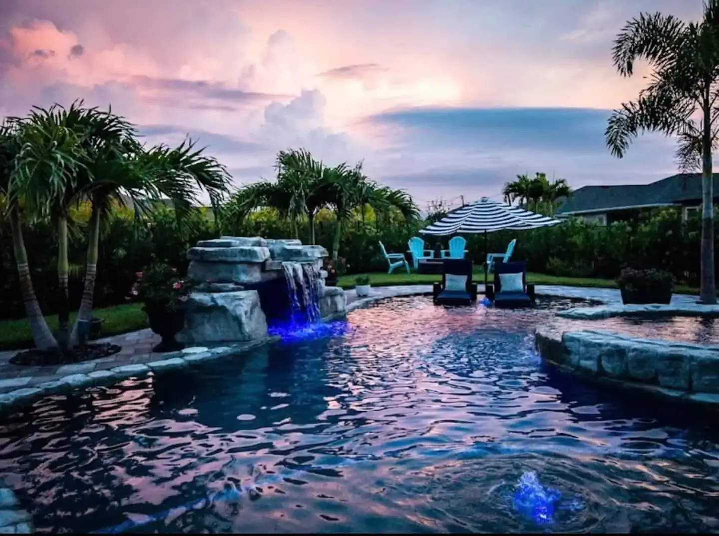 Twilight resort pool with blue-lit water, waterfalls, loungers, and palm trees under a pink sky